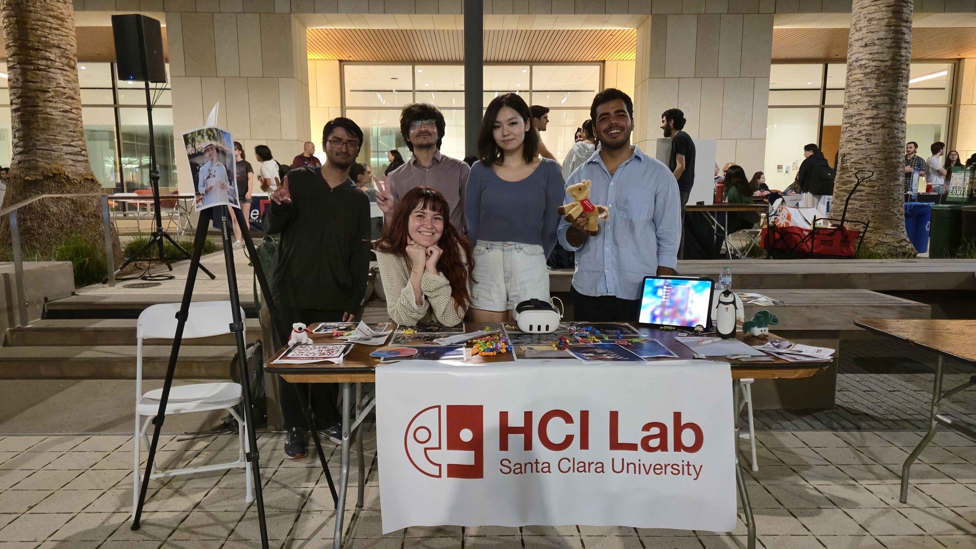 Five people behind an HCI Lab demo table with a VR headset, tablet, and printed materials.