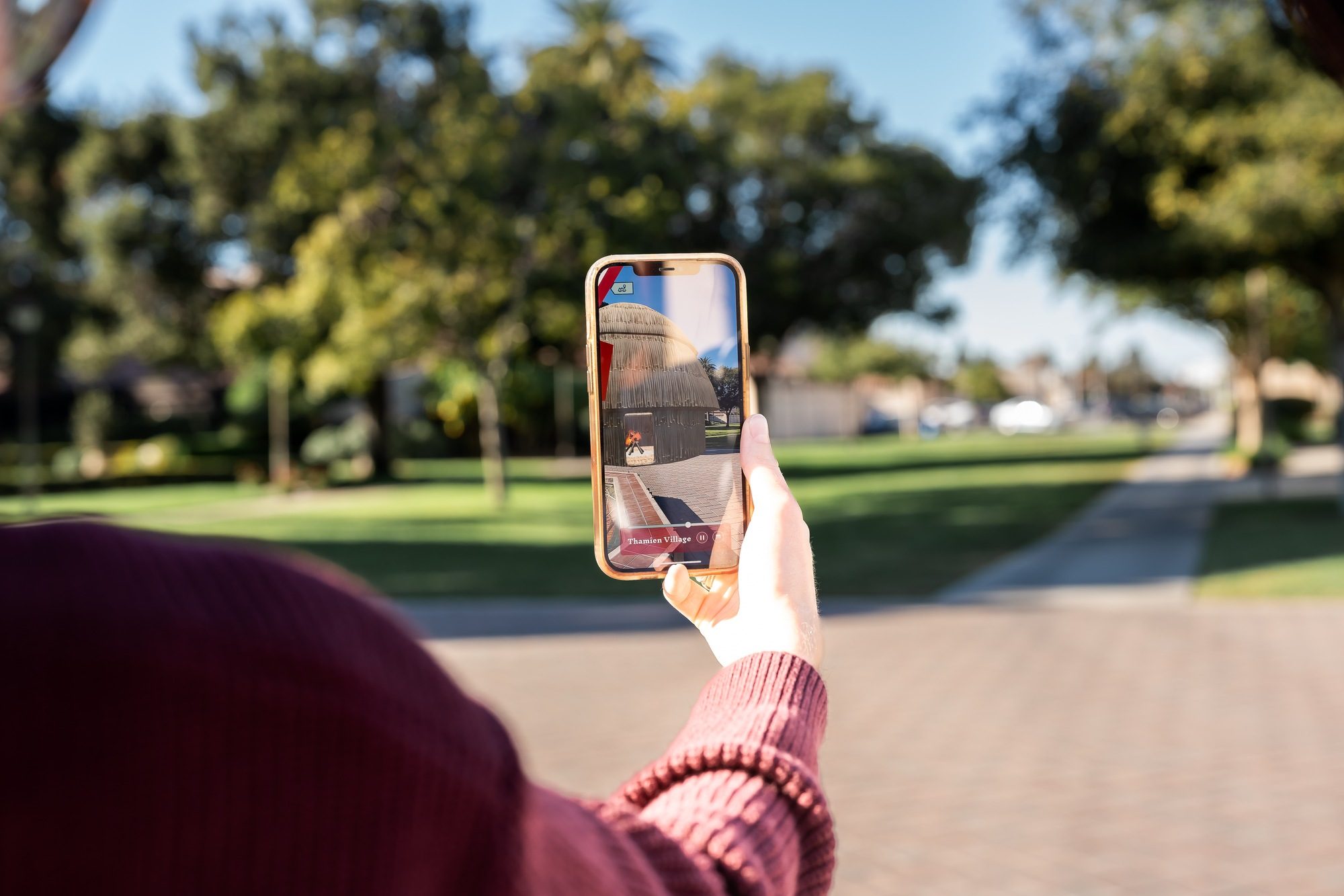 A visitor holds a phone displaying an augmented-reality tule dwelling at the Thámien Village stop of the Ohlone AR Tour.