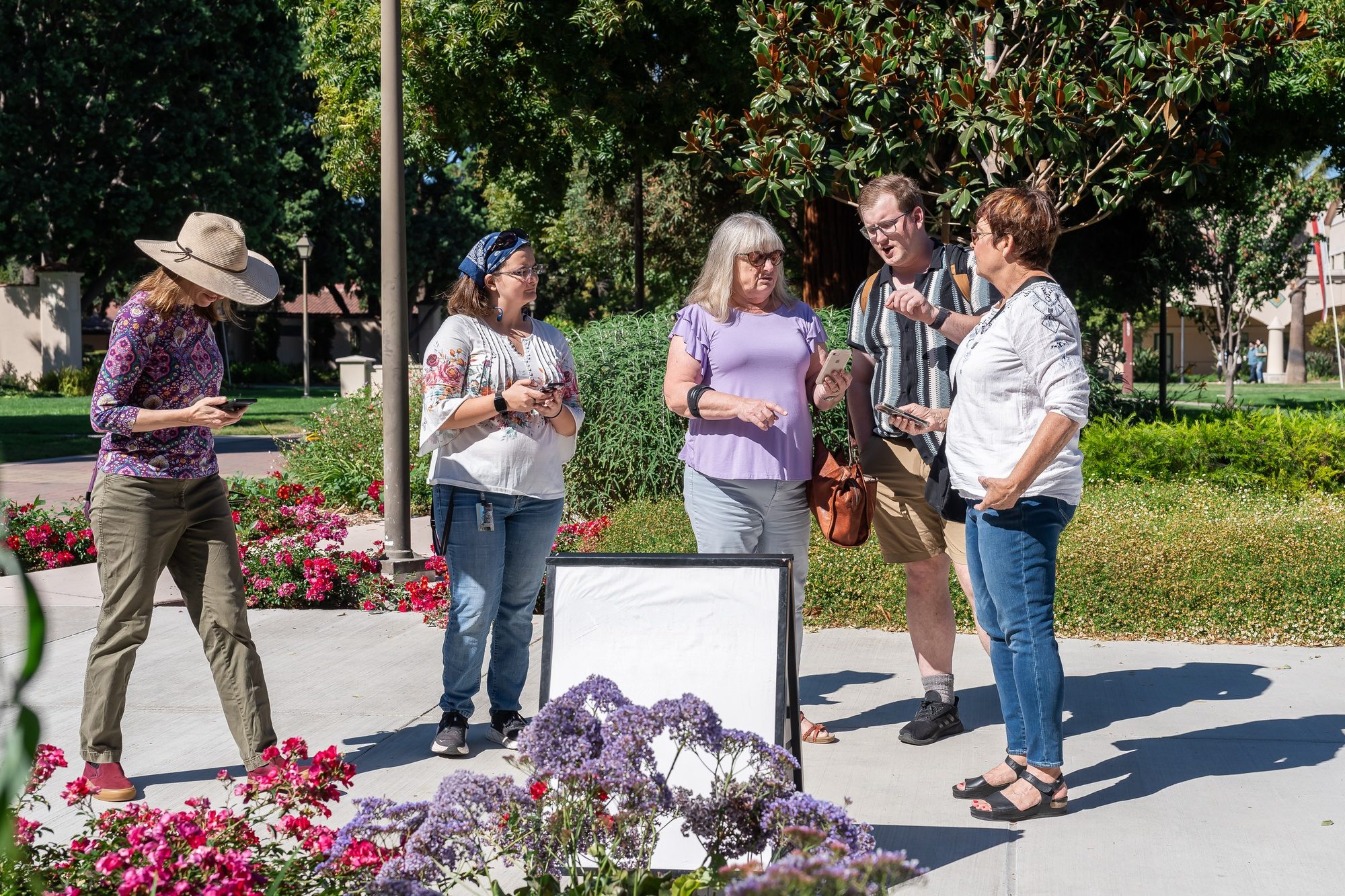 Five visitors stand among flowers at Santa Clara University, holding phones and discussing a tour stop.